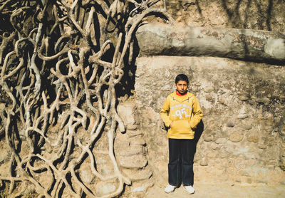 Portrait of boy with hands in pockets standing by tree roots against wall at old ruins