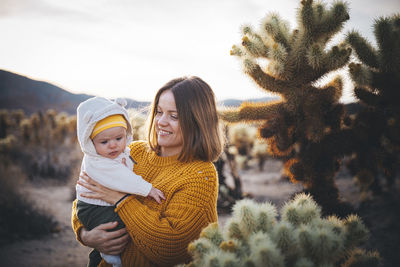 A woman with a baby is standing near a cactus in the desert