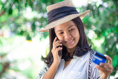 Mid adult woman with credit card talking over mobile phone while standing against trees