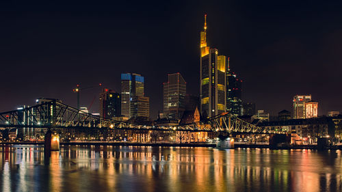 Illuminated bridge over river by buildings against sky at night