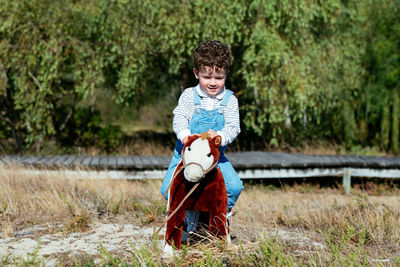 Full body of adorable curly boy in denim overall sitting on soft toy horse and riding while having fun in countryside field