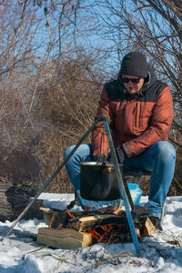 Man by campfire, pot of soot over bonfire hanging on tripod, winter outdoor cooking at campsite