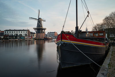 Boats moored at harbor in river by windmill against sky