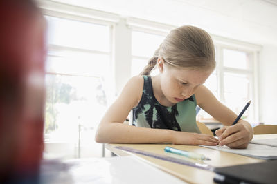 Girl writing in book against window at classroom