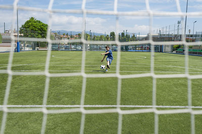 Boy kicking soccer ball against sky seen through net