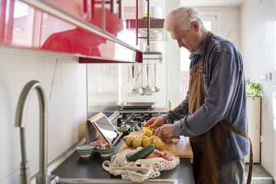 Senior man chopping raw potatoes on cutting board in kitchen