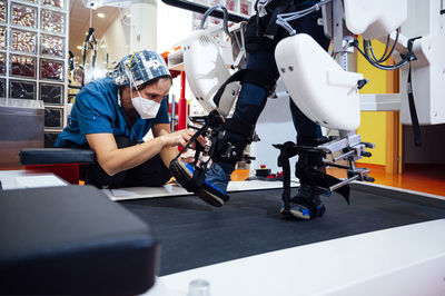 Female specialist in protective mask fastening belts on legs of unrecognizable patient with disability while preparing for physiotherapy exercise on modern equipment in rehabilitation center