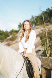 Young long hair woman in white shirt riding white horse on seascape background