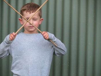 Boy holding sticks while standing against corrugated iron