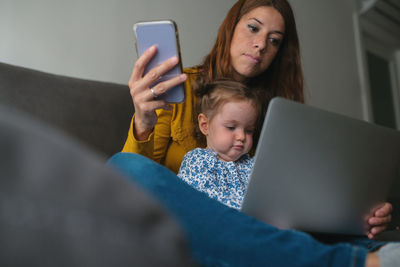 From below young female freelancer using mobile phone while working with laptop on sofa in living room with little daughter