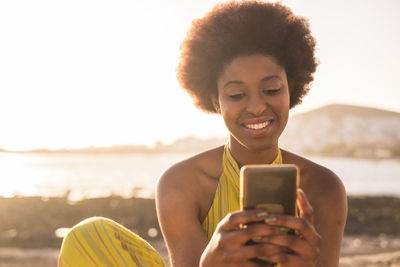 Smiling young woman using smart phone against sky during sunset