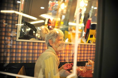 Cheerful senior woman looking away while sitting in restaurant seen through window