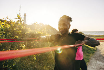 Portrait of smiling man pulling resistance band while exercising at beach