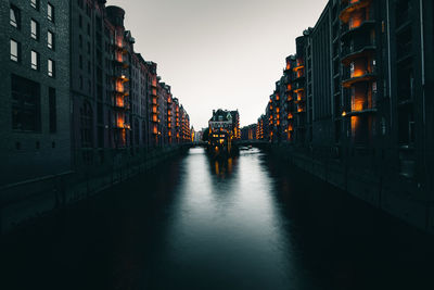 Illuminated street amidst buildings against sky at dusk