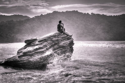 Side view of man sitting on rock formation in sea against mountains