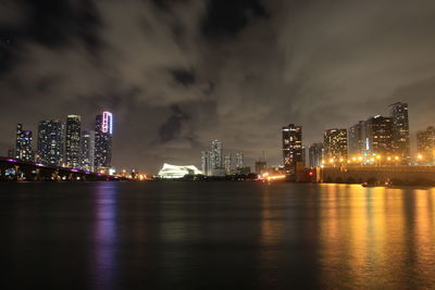 Illuminated modern buildings by sea against sky at night