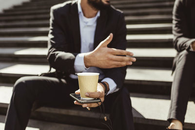Midsection of businessman with disposable cup making hand gesture while sitting on staircase