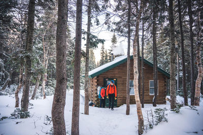 Two men with jackets and backpacks exit a cabin in the a snowy forest