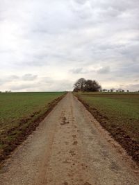Dirt road amidst fields against cloudy sky