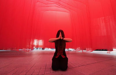 Woman with hands clasped kneeling on floor against red curtain