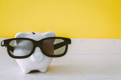 Close-up of sunglasses with piggy bank on table against yellow wall