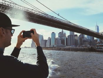 Low angle view of man photographing bridge with city in background