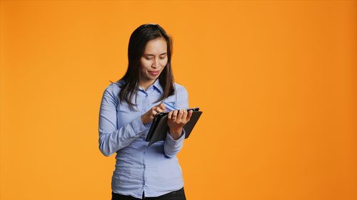 Young woman using mobile phone while standing against yellow background