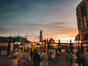 People walking on illuminated building in city against sky during sunset