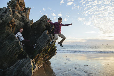 Playful boy jumping at beach while brother sitting on rocks