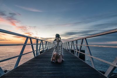 Man sitting on pier over sea against sky during sunset