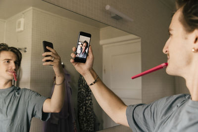 Man brushing teeth taking selfie through mobile phone in bathroom at home