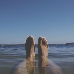 Low section of man with feet up in sea against clear sky