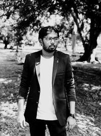 Young man wearing blazer and eyeglasses looking away while standing in park