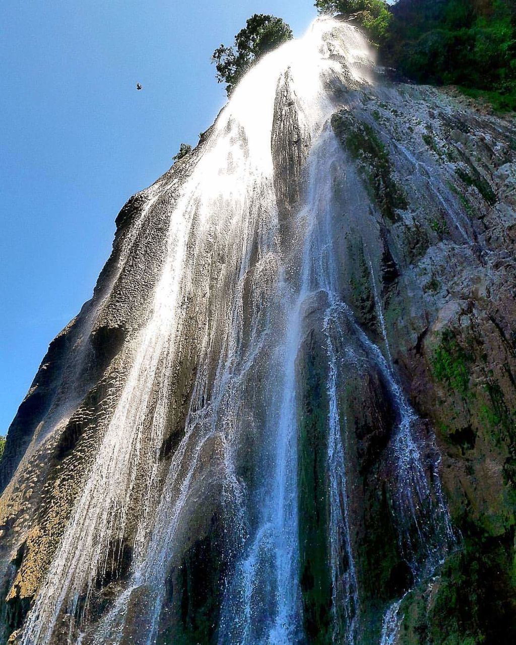 motion, low angle view, blue, spraying, clear sky, fountain, water, beauty in nature, sunny, splashing, nature, day, outdoors, power in nature, flowing water, mountain, scenics, sky, sun, flowing, majestic, multi colored, waterfront, mountain range