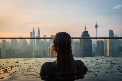 Rear view of woman swimming in infinity pool against city