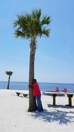 Full length of man hugging tree at beach against clear blue sky