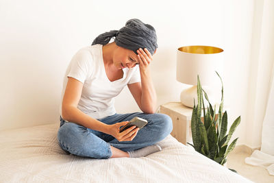 A middle-aged woman sits on a bed looking at a smartphone screen.