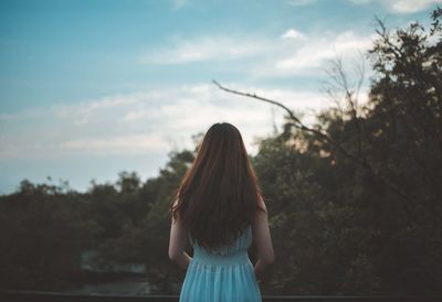 Rear view of woman standing by trees against cloudy sky