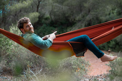 Portrait of man sitting using smart phone while on hammock in forest