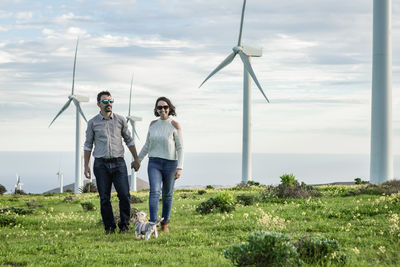 Couple holding hands while walking on grass against wind turbines