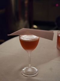 Paper plane cocktail in a cocktail glass on a white clothed table, garnished with an origami plane