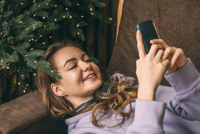 Beautiful woman lies near decorated christmas tree on sofa, using phone, typing message, new year