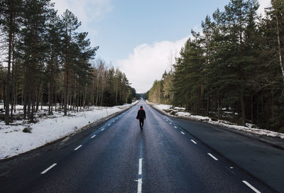 Rear view of young man walking on road amidst trees against sky in winter