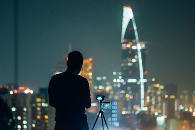 Silhouette man standing by digital camera on tripod against illuminated cityscape at night