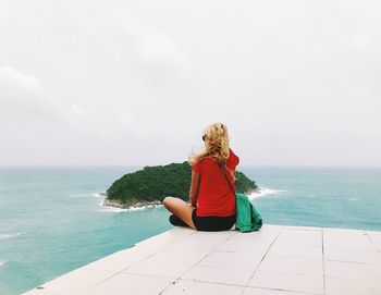 Rear view of mid adult woman looking at sea while sitting on tile against sky