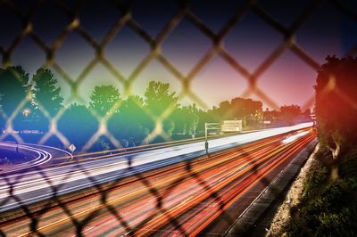 Light trails on road in city against sky seen through chainlink fence