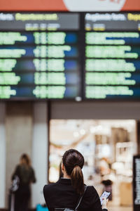 Rear view of businesswoman holding smart phone while looking at arrival departure board at railroad station