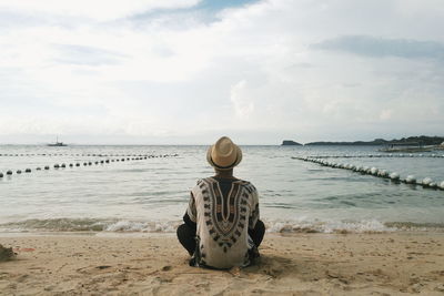 Rear view of man sitting at beach against cloudy sky