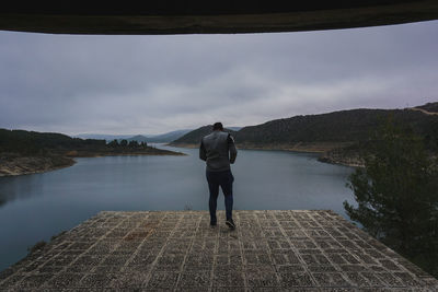 Rear view of man walking at observation point by river against cloudy sky