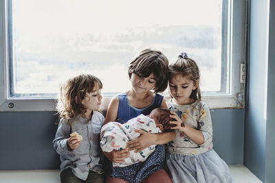 Front view of three siblings holding newborn brother against window